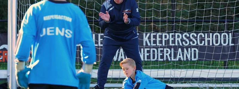 Keeperstrainer moedigt jonge keeper aan tijdens een trainingssessie bij de Keepersschool van Keepersaction. De focus ligt op het verbeteren van de vaardigheden van de jeugdkeeper, die geconcentreerd de bal vastpakt, met een andere jonge keeper op de voorgrond en het doelnet op de achtergrond.